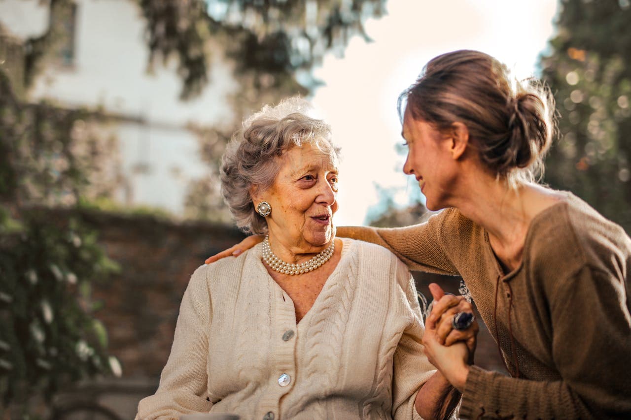 An older woman and her adult daughter holding hands and smiling together outdoors.